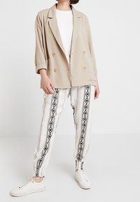 Osoba v béžovém dvouřadovém blejzru, bílém topu, bílých kalhotách s černými geometrickými pruhy a bílých teniskách postávající na bílém pozadí.