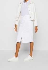 Falda de denim blanca con abertura frontal, combinada con una camiseta polo azul clara y una chaqueta de denim blanca. La modelo lleva zapatillas blancas.
