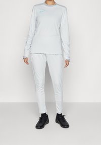 Světle šedá dlouhá tričko s dlouhým rukávem a odpovídající kalhoty, vyrobené z hladkého materiálu, s akcenty tyrkysového loga Nike a přiléhavým designem. Černé sportovní boty.