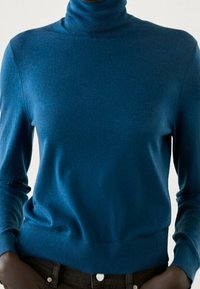 Person wearing a fitted blue turtleneck sweater with long sleeves and dark pants, standing against a neutral background.