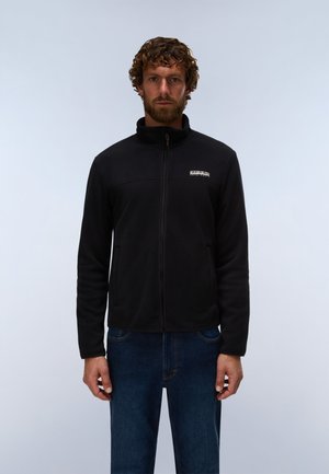 Mann mit lockigem Haar und Bart trägt eine schwarze Jacke mit Reißverschluss und blaue Jeans und steht vor hellem, einfarbigem Hintergrund.