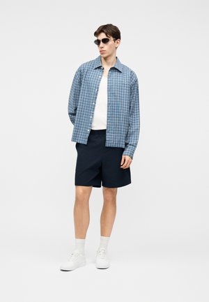 Ung mand iført sorte solbriller, blå ternet skjorte med knapper over en hvid t-shirt, sorte shorts, hvide sokker og hvide sneakers.
