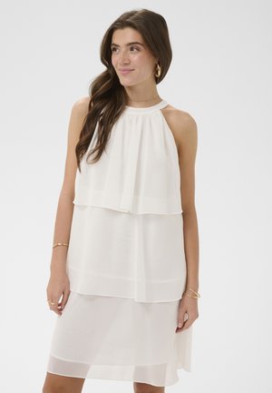 Ung kvinde med langt brunt hår iført hvid, ærmeløs lagdelt kjole samt guldringe, armbånd og øreringe mod ensfarvet baggrund.