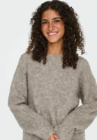 Jeune femme aux cheveux bruns bouclés et avec des taches de rousseur souriante, portant un pull en maille gris clair et les mains jointes devant elle.