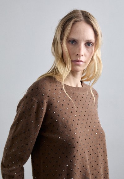 Blondynka z luźnymi falami, ubrana w brązowy sweter ozdobiony małymi metalowymi ćwiekami, patrząca bezpośrednio w kamerę na jednolitym tle.