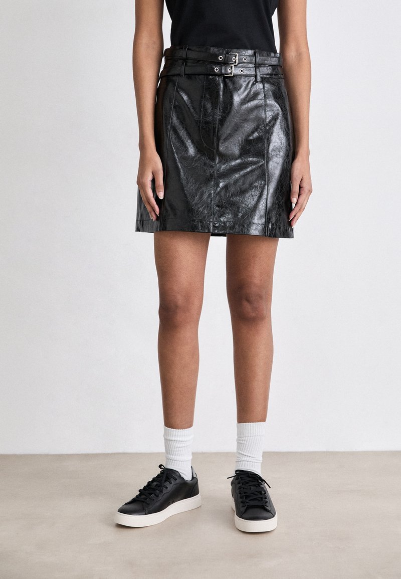 Gonna mini in pelle nera con vita con cintura, cuciture dettagliate e una texture lucida, abbinata a sneakers nere e calze bianche.