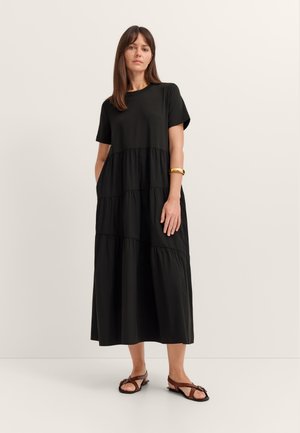 Frau steht und trägt ein schwarzes, gestuftes Midi-Kleid mit kurzen Ärmeln, braune flache Sandalen und ein goldenes Armband am Arm.
