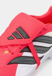 Φωτεινό ροζ ποδοσφαιρικό παπούτσι με υφή στην πάνω πλευρά, μαύρες λεπτομέρειες και λευκή σόλα, με χαρακτηριστικό λογότυπο τριών ριγών και λεπτομέρεια από ελαστική ταινία.