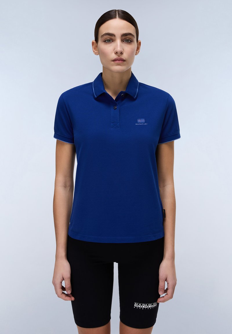 Mörkblå pikétröja med krage och korta ärmar. Har en liten logga på vänster bröst och kontrasterande sömmar. Bärs med svarta shorts.