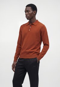 Homme portant un polo à manches longues couleur rouille et un pantalon noir, les mains dans les poches, debout devant un fond clair uni.
