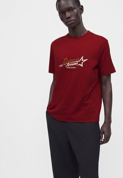 Ενήλικος άνδρας που φοράει κόκκινο T-shirt Emporio Armani με σχέδιο αστέρι και μαύρο παντελόνι, στέκεται μπροστά σε απλό ανοιχτό φόντο.