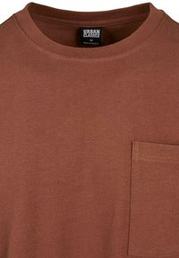 Camiseta de algodón marrón con cuello redondo, que presenta un bolsillo en el pecho izquierdo y un acabado suave y texturizado. Etiqueta de la marca visible en el escote.