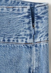 Gros plan sur un tissu en denim bleu avec une fermeture éclair bleue partiellement ouverte et des coutures orange visibles.