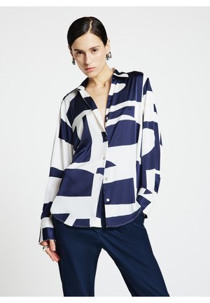 Chemise blanche soyeuse avec des motifs géométriques abstraits bleu marine, boutonnée sur le devant, à manches longues, associée à un pantalon bleu marine.