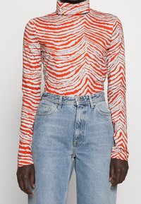 Långärmad polotröja i orange och vit vågmönster, tillverkad av stretchigt tyg, ihop med ljusblå jeans med hög midja.