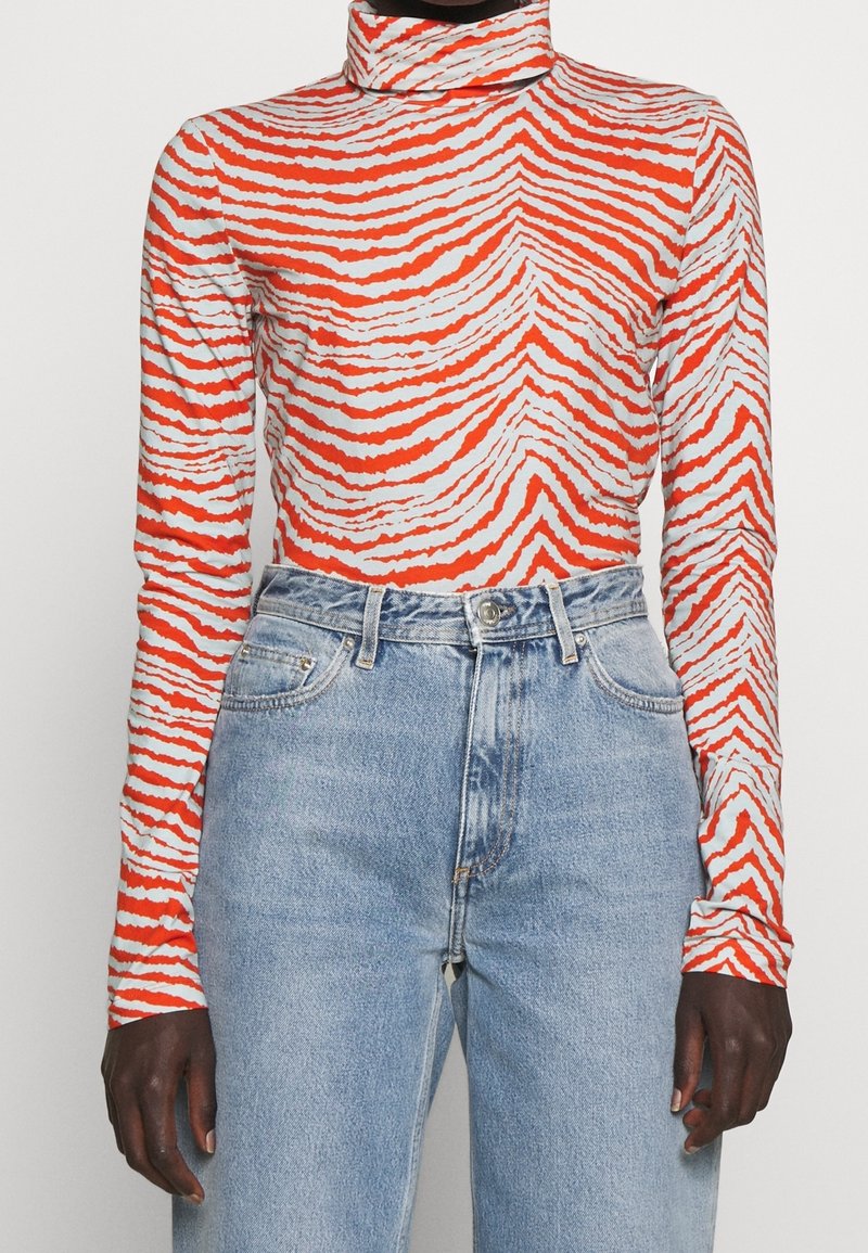 Långärmad polotröja i orange och vit vågmönster, tillverkad av stretchigt tyg, ihop med ljusblå jeans med hög midja.