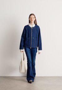 Τζιν μπουφάν σε σκούρο μπλε με φερμουάρ μπροστά, δύο τσέπες και στρογγυλεμένο ντεκολτέ. Συνδυάζεται με τζιν με καμπάνα και χρωματιστή τσάντα ώμου σε εκρού.