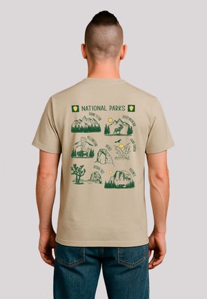Beige short-sleeve t-shirt featuring green illustrations of various national parks, with names and scenic elements arranged on the back.
