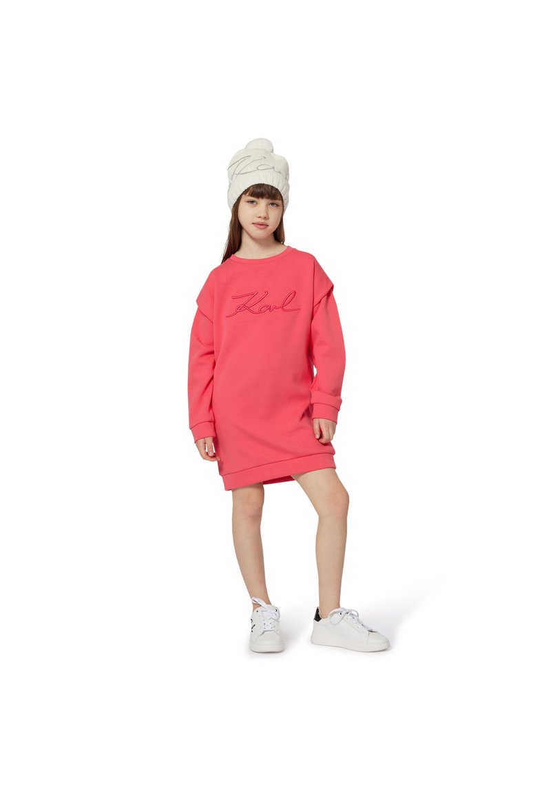 Rosa sweatshirt-kjole med oversized passform, ribbestrikkede mansjetter og kant. Har "Karl" brodert i kursiv. Hvit strikket lue og hvite joggesko.