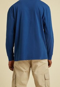 Langärmliges, blaues Baumwoll-T-Shirt mit Rundhalsausschnitt, von hinten gezeigt. Getragen mit hellbeigen Cargo-Hosen. Glatte Textur.