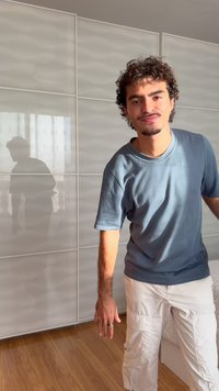 Νέος άνδρας με σγουρά μαλλιά που φοράει μπλε κοντομάνικη μπλούζα και λευκά cargo σορτς, στέκεται σε εσωτερικό χώρο κοντά σε μια ντουλάπα με φως που αντανακλά πάνω του.