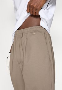 Pantaloni bej cu talie elastică, dotati cu buzunare laterale și un detaliu subtil al logo-ului. Țesătura pare netedă, cu un ușor luciu.