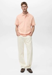 Svetle růžová polo košile, volný střih, krátké rukávy, zkombinovaná se krémovými kalhotami. Hnědé boty dotvářejí outfit. Jednoduchý design, žádné vzory.