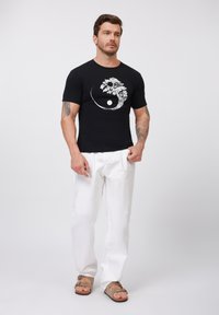 Black t-shirt featuring a white tree and yin-yang graphic, paired with white trousers and brown sandals. Smooth fabric, fitted design.
