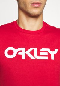 Czerwona koszulka wykonana z bawełny, z wyrazistym białym logo "OAKLEY" w nowoczesnej czcionce. Okrągły dekolt i krótkie rękawy.