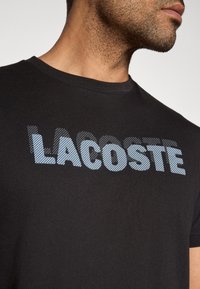 Czarna koszulka z logo "LACOSTE" w jasnoniebieskie kropkowane litery. Zbliżenie podkreśla miękkość tkaniny i okrągły dekolt.