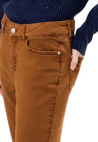 Pantalones de mezclilla marrones con una textura suave, que cuentan con bolsillos delanteros y un cierre de botón plateado en la cintura.