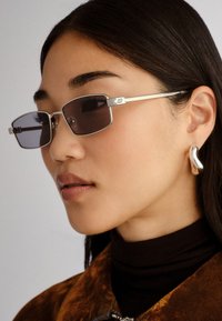 Ochelari de soare cu ramă pătrată și lentile închise, cu accente metalice argintii și un design minimalist, purtați cu un pulover negru cu guler înalt și o jachetă texturată.