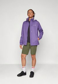 Purpurinė neperšlampama striukė su kapišonu, derinama su juoda marškinėliais ir žaliais šortais. Modelis dėvi juodus batus ir dryžuotas baltas kojines.