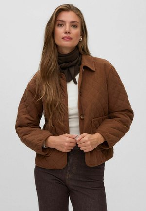 Vrouw met lang haar die een bruine doorgestikte jas draagt, een donkere sjaal, een wit top en donkere broek, kijkt naar voren en past haar jas met haar handen aan.