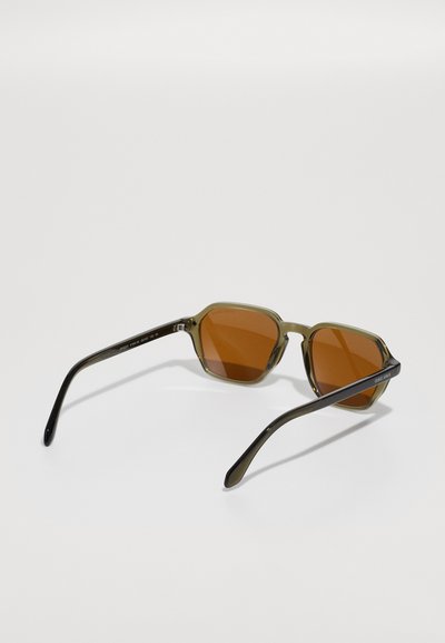 Brown-tinted sunglasses with an octagonal frame. Transparent olive green plastic material. Slim black arms, minimal branding detail on the side.