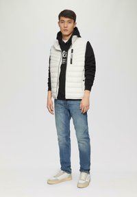 Witte gewatteerde mouwloze vest over een zwarte hoodie, gecombineerd met blauwe jeans en witte sneakers met beige accenten. Effen achtergrond.