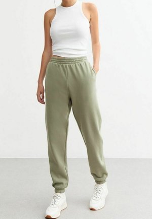 Lightweight white tank top paired with olive green sweatpants featuring an elastic waistband and cuffed ankles. The outfit is completed with white sneakers.