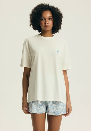 Žena s kučeravými vlasmi nosiaci voľné biele tričko s malým modrým vzorom a svetlomodré šortky s bielymi listovými vzormi.