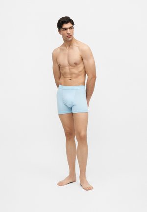 Ung mand står barfodet iført lyseblå boxershorts og kigger til siden mod en ensfarvet lys baggrund.