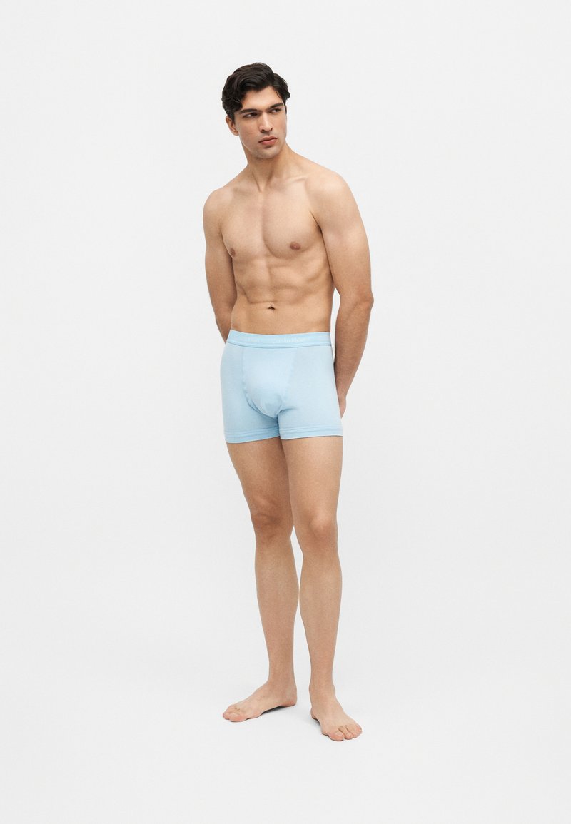 Jeune homme debout pieds nus, portant un boxer bleu clair, regardant sur le côté sur un fond clair uni.