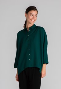Camicia verde con bottoni, dalla vestibilità comoda, maniche lunghe e orlo asimmetrico. Realizzata in tessuto morbido e liscio con dettaglio del colletto.