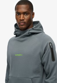 Sudadera con capucha gris de textura suave, con un logo verde en el pecho y un bolsillo con cremallera en la manga.