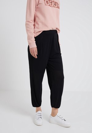 Personne portant un pantalon noir fuselé avec des poignets élastiques, un haut rose à manches longues et des baskets blanches, debout devant un fond uni.