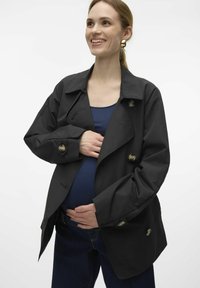 Svart trenchcoat med stora knappar, överdimensionerad passform, sidofickor och en mjuk textur, kombinerad med en mörkblå topp och jeans.