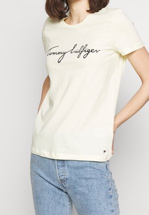 Kvinde iført lysegul kortærmet T-shirt med "Tommy Hilfiger" i sort skrift og lille logo nær kanten, kombineret med blå jeans.