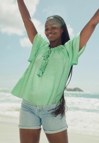 Blusa verde chiaro a maniche corte con scollatura arricciata e dettaglio a fiocco, abbinata a pantaloni corti in denim, su uno sfondo di spiaggia.