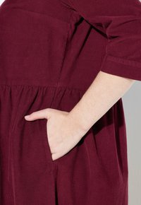 Käsi burgundinpunaisessa corduroydressissä, jossa on kootut vyötärö ja kolme neljännestä hihoista.