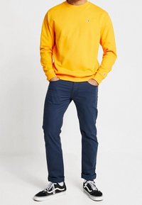 Felpa in cotone arancione, pantaloni blu navy e sneakers nere con dettagli bianchi; vestibilità casual con polsini a coste e scollatura rotonda.