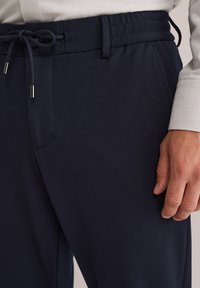 Temno modre hlače z teksturirano tkanino, z elastičnim pasom in vrvico, stranskimi žepi ter elegantnim ozkim krojem.