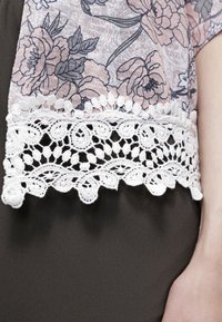 Durchsichtiger Stoff mit floralen Mustern in hellrosa und blauen Tönen, versehen mit einem weißen Spitzenrand mit filigranen Details auf einem dunklen Stoffhintergrund.
